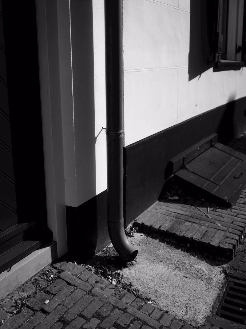 A metal downspout turning out at the base of a building wall and discharging directly onto the paving beside the foundation